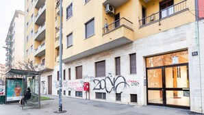 A multi-story building with a bus stop, graffiti on the wall, and a storefront with glass doors. - GuestHost - Lovely studio apartment complete with all major amenities (Milano)