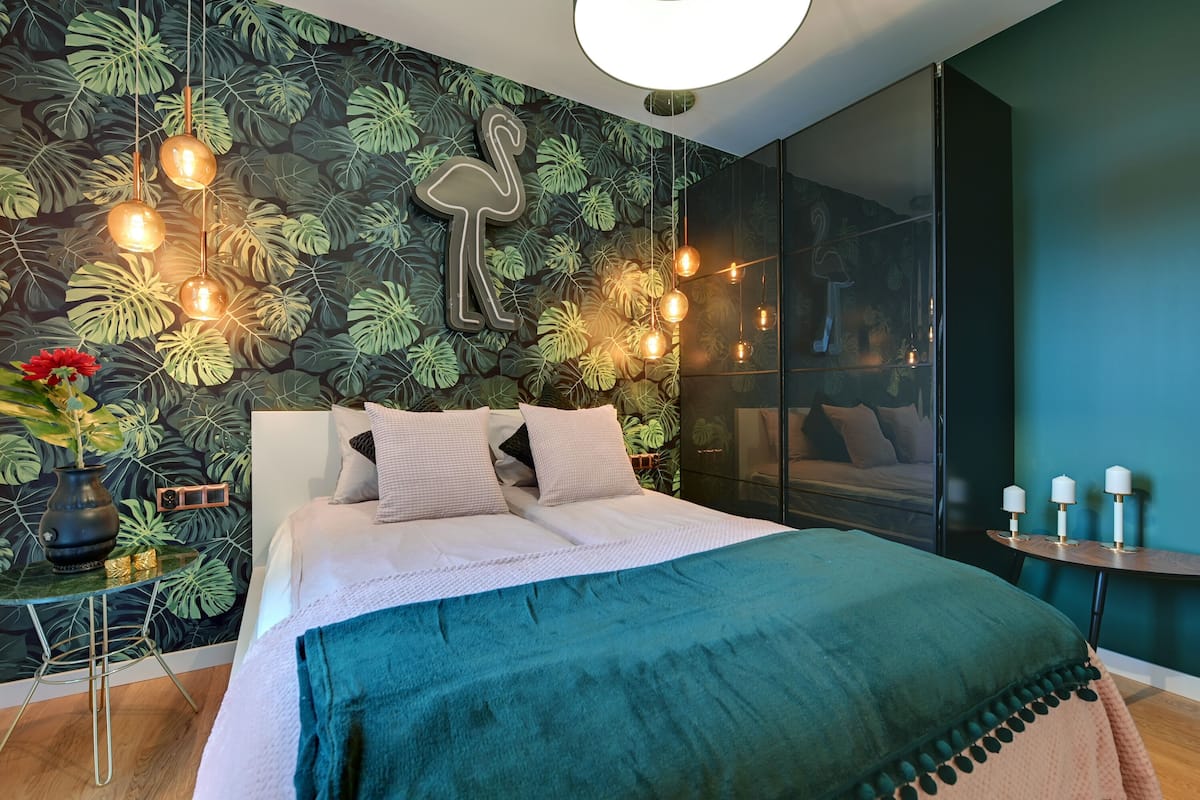A modern bedroom with a large bed, a wall with tropical leaf wallpaper, hanging pendant lights, and a bedside table with candles.