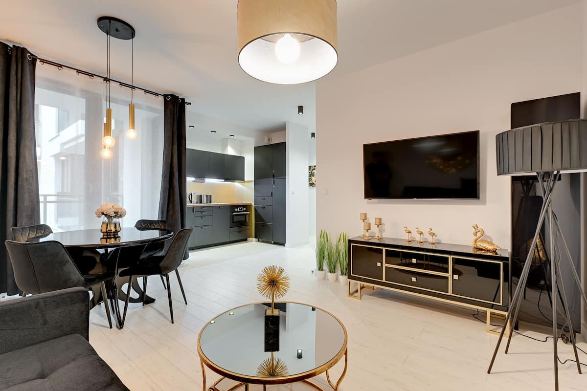A modern living room with a glass coffee table, black and gold furniture, a large flat-screen TV, and a dining area with a black table and chairs.