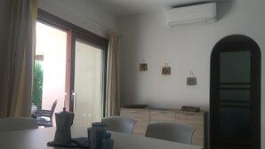 Een eethoek met een tafel, stoelen en een koffiezetapparaat. Er is een raam met gordijnen, een deur en een wandairconditioner.