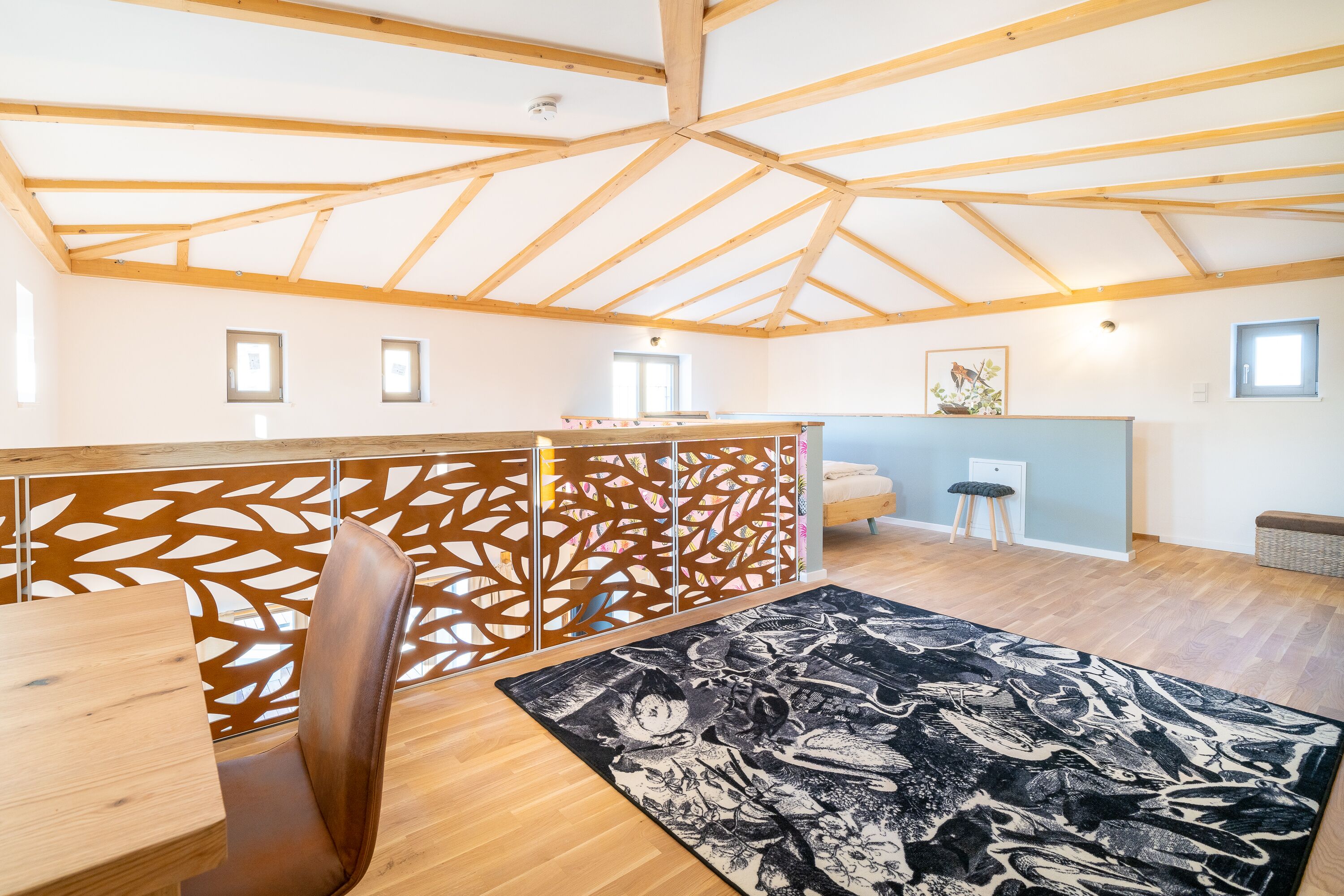 A spacious room with a high ceiling and wooden beams, a dining area with a table and chairs, a decorative rug, and a view of the outside through a window.