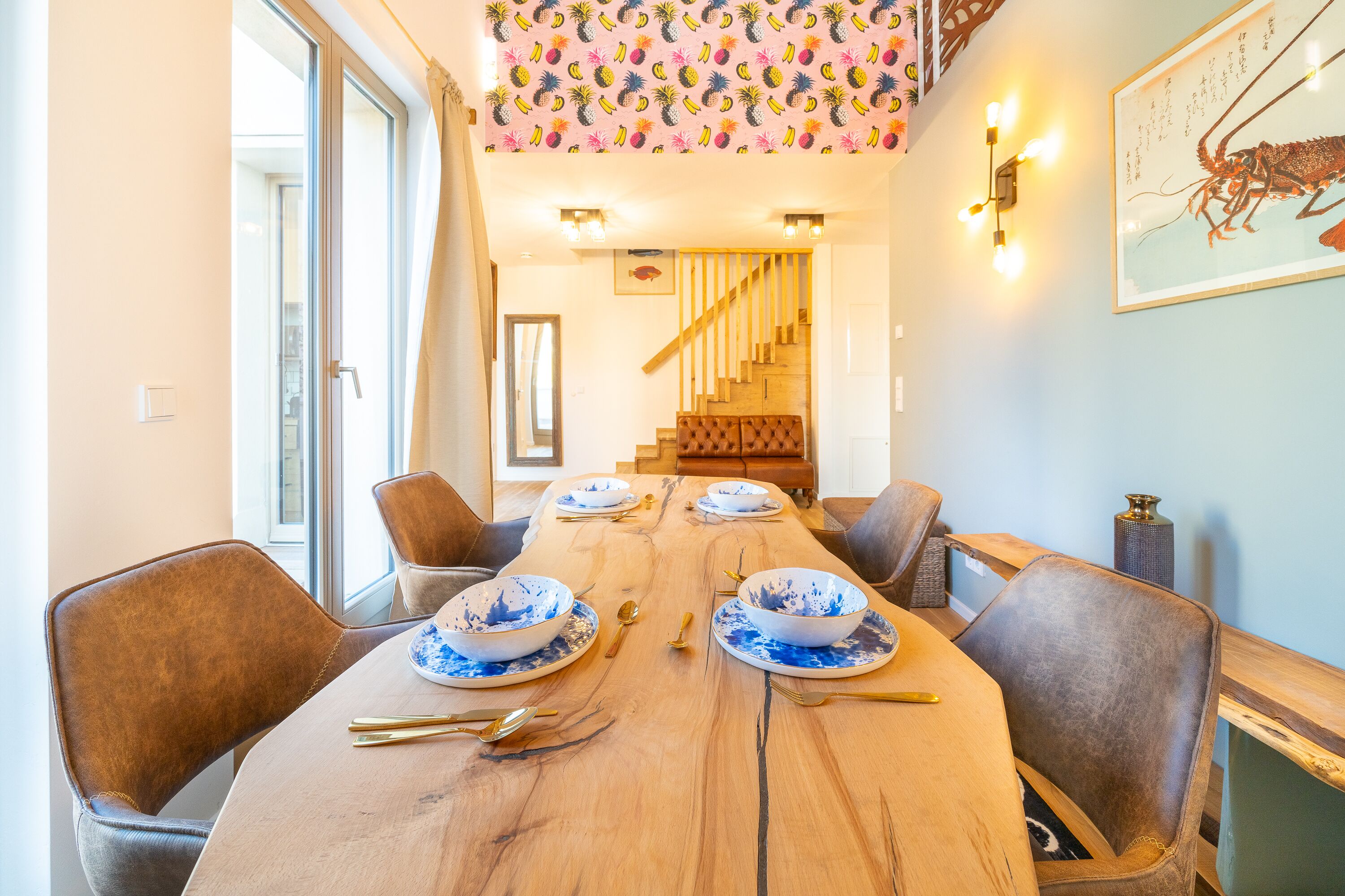 A dining area with a wooden table set for four, surrounded by leather chairs. A staircase is visible in the background leading to an upper level.