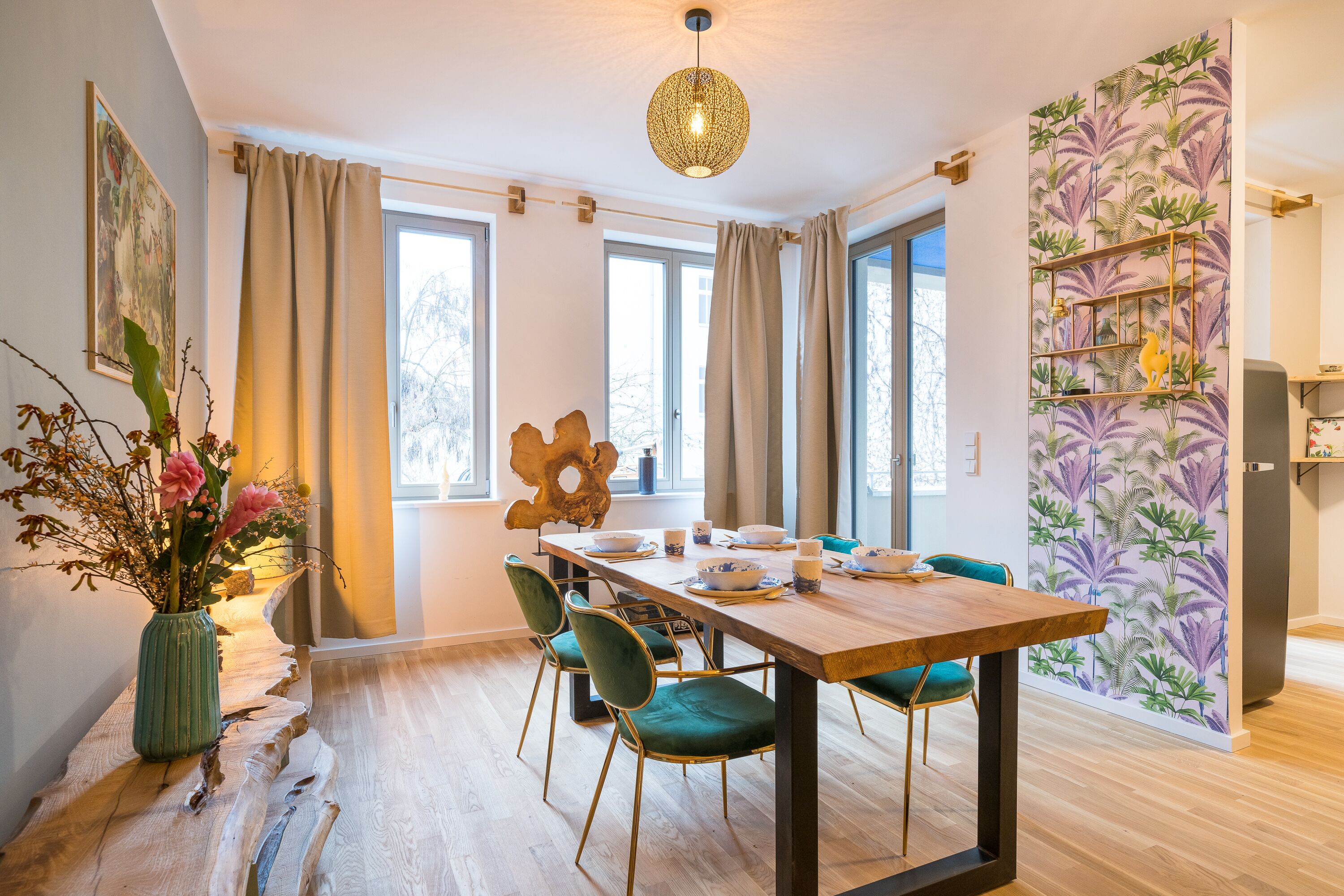 A dining area with a wooden table, green chairs, a vase with flowers, and a wall with a tropical wallpaper.