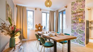 A dining area with a wooden table, green chairs, a vase with flowers, and a wall with a tropical wallpaper.