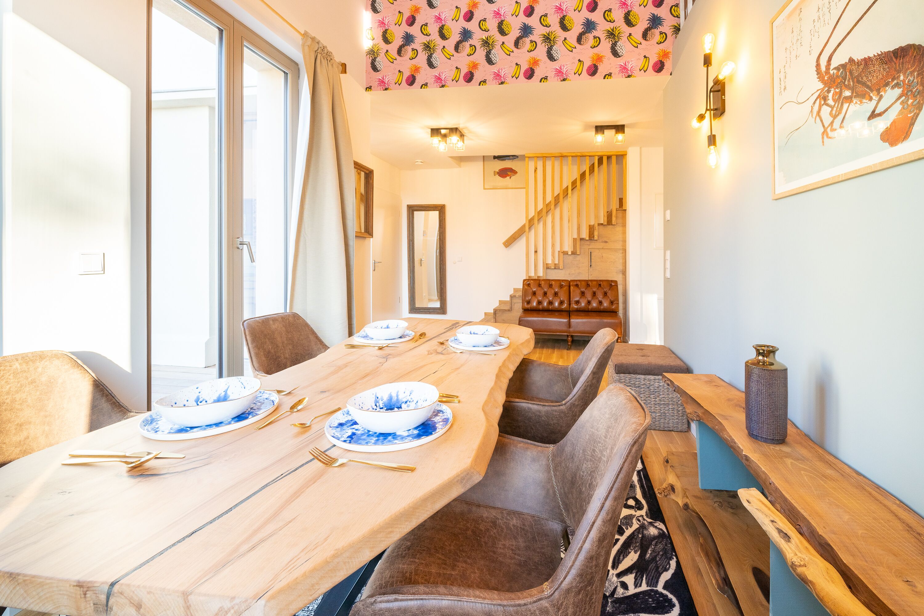Un comedor con una larga mesa de madera dispuesta para cuatro personas, rodeada de sillas acolchadas. Se ve una escalera con peldaños y barandilla de madera al fondo.
