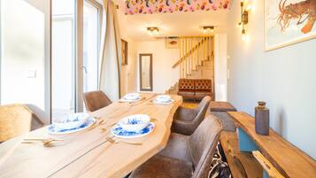 Un comedor con una larga mesa de madera dispuesta para cuatro personas, rodeada de sillas acolchadas. Se ve una escalera con peldaños y barandilla de madera al fondo.