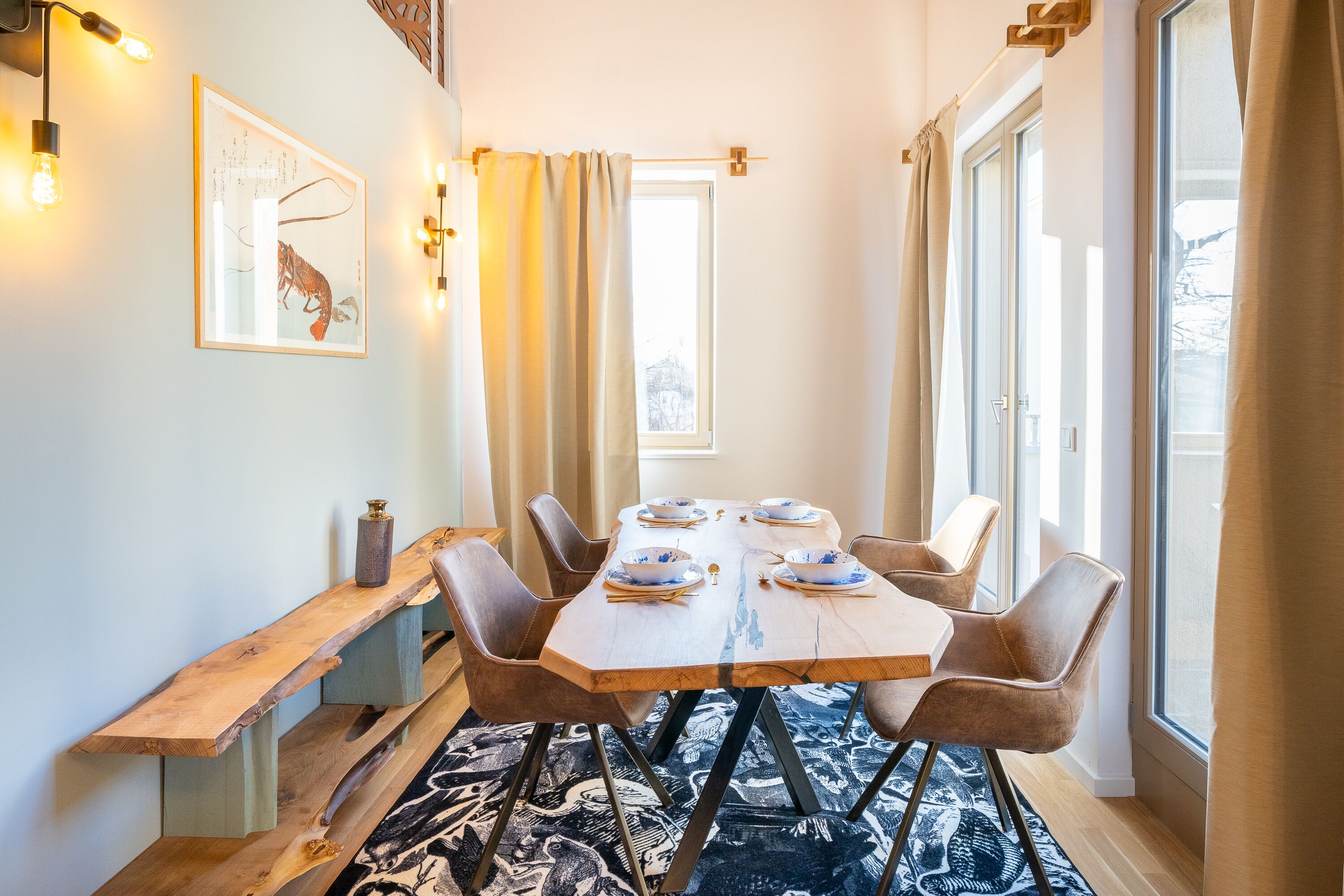 Un coin repas avec une table en bois, six chaises et un banc. Les fenêtres sont dotées de rideaux, un luminaire est fixé au mur et un tableau encadré orne le mur.