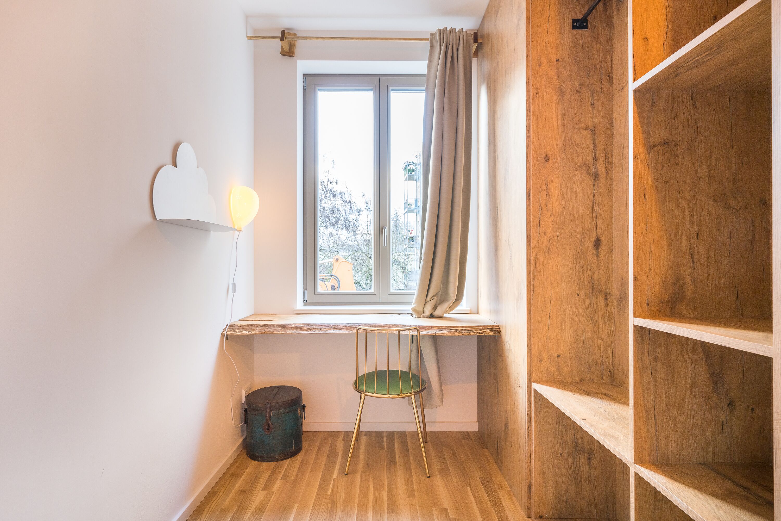 Une pièce d’angle avec un plancher en bois, une fenêtre avec des rideaux, un fauteuil vert et une étagère.