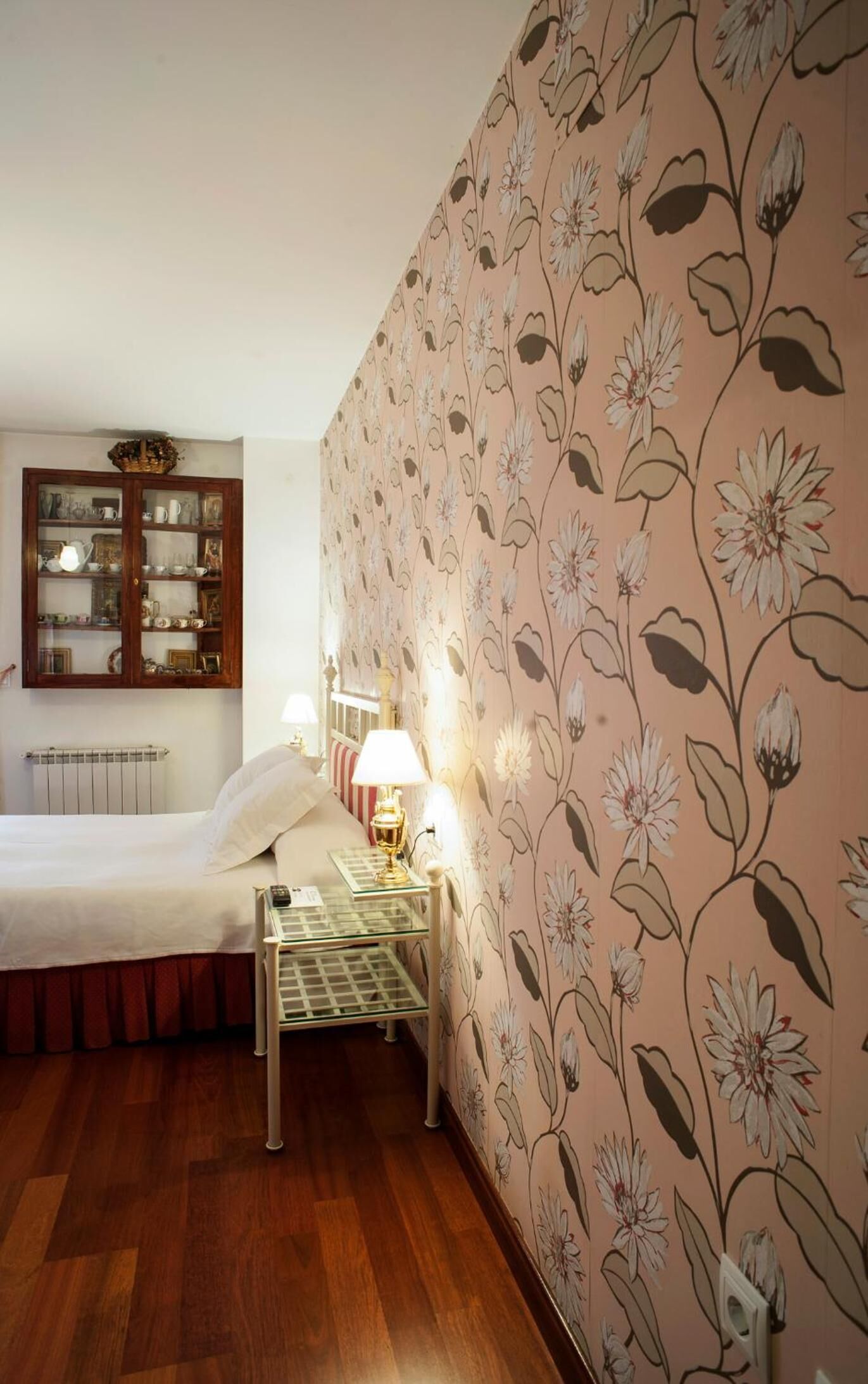 Una camera da letto con un armadio in legno, un letto con cuscino a righe rosse e bianche, un comodino con una lampada e carta da parati con motivi floreali.