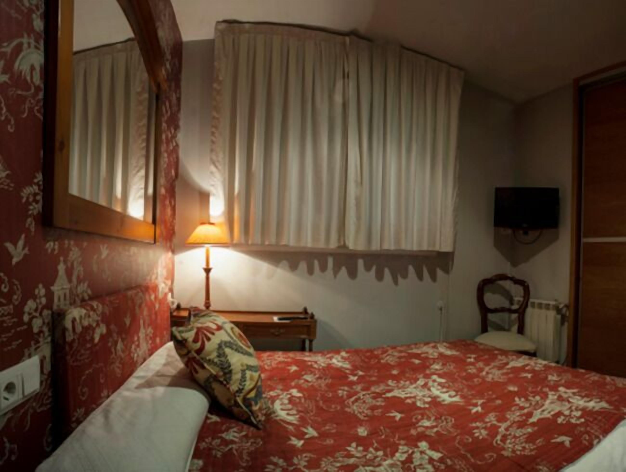 A bedroom with a bed, a nightstand, a lamp, a mirror, and a TV mounted on the wall.