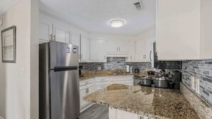 Cozinha moderna com armários brancos, geladeira de aço inoxidável, bancadas de granito e painel de azulejos.