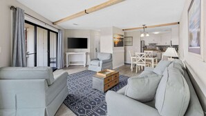 Uma sala de estar moderna com um sofá cinzento, uma mesa de centro e uma televisão. Há uma área de jantar com uma mesa e cadeiras. A cozinha é visível ao fundo, com armários brancos e uma bancada.