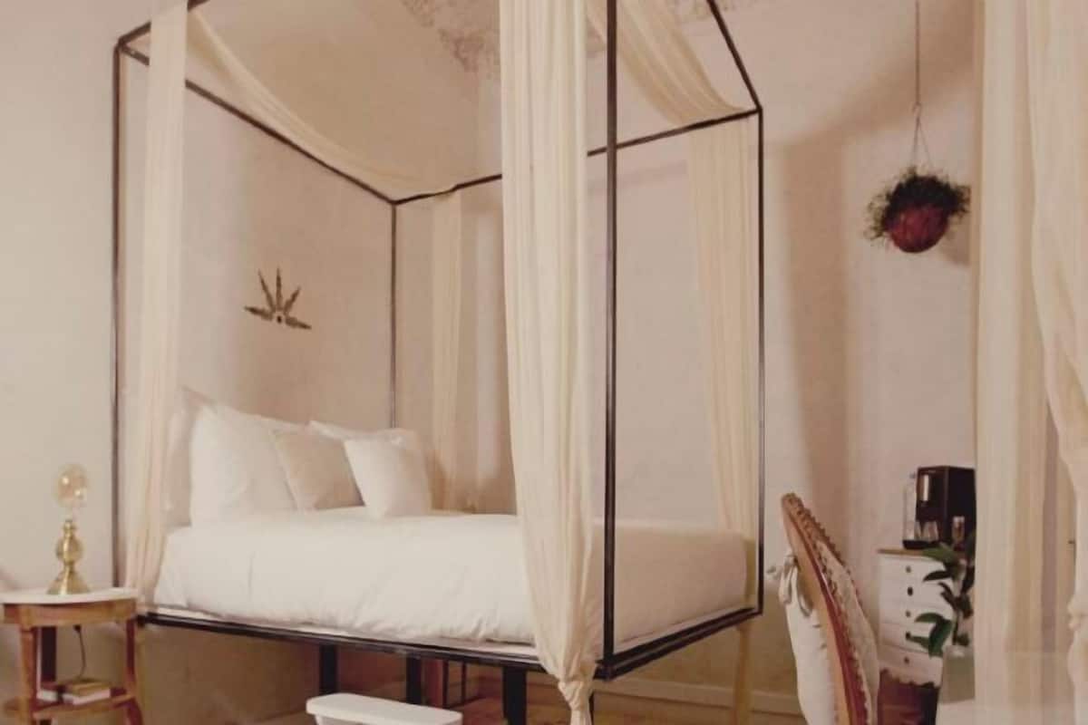 A four-poster canopy bed with a white bedspread and pillows, a bedside table with a lamp, and a chair with a decorative backrest.
