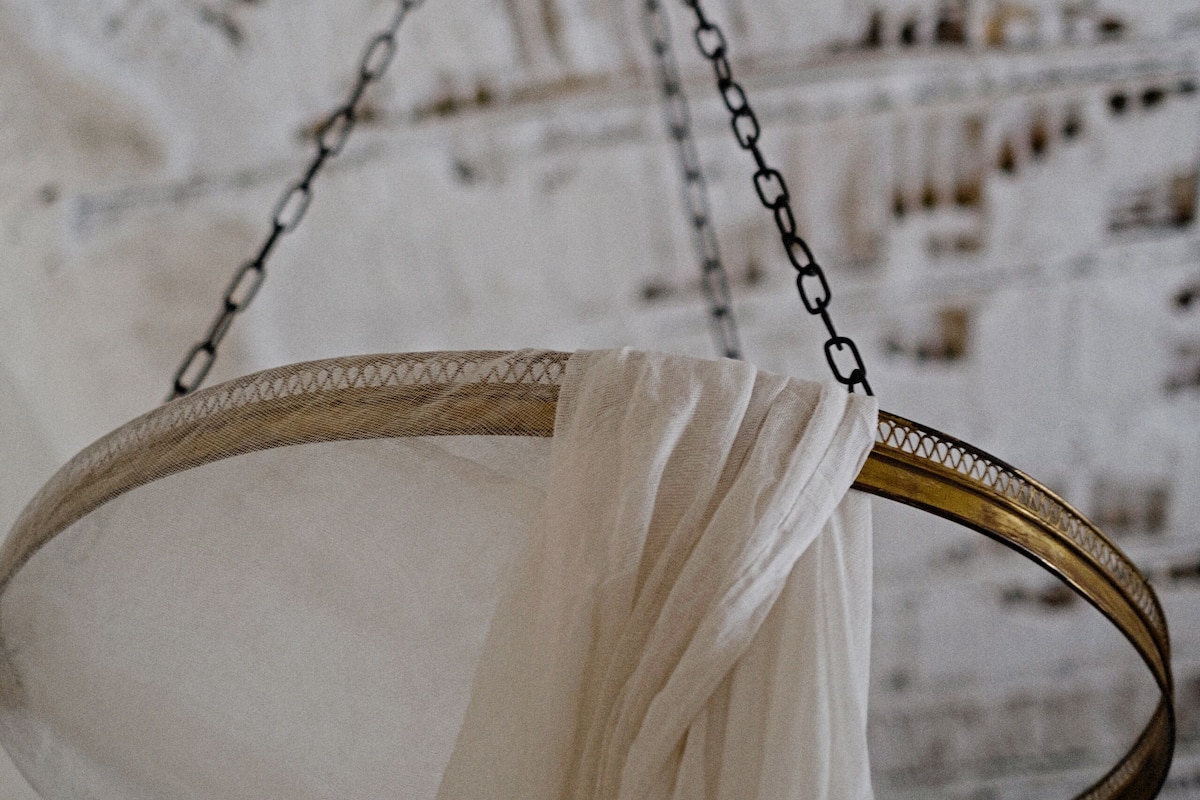 A white, ornate mirror with a chain hanging from its center, set against a textured, white wall.