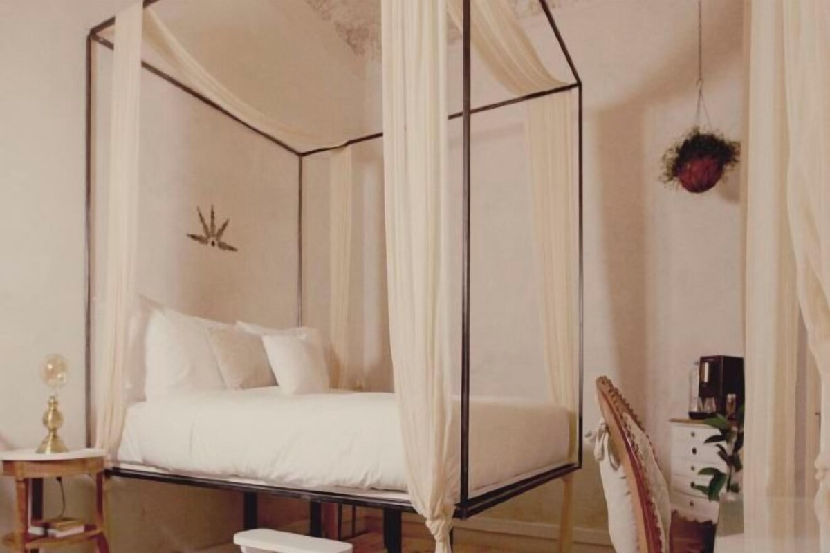 A four-poster canopy bed with a white bedspread and pillows, a bedside table with a lamp, and a chair with a decorative backrest.