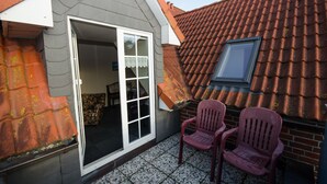 Een kleine kamer met een betegelde vloer, een raam en een deur naar een buitenruimte met twee stoelen en een rood dak.