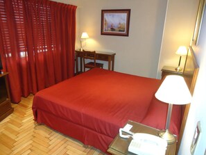 A hotel room with a large bed, a desk with a lamp, a chair, a telephone, and a painting on the wall. - Gran Hotel Mercedes (Mercedes)
