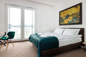 Une chambre d’hôtel avec un grand lit, un fauteuil vert, un tableau au mur et une vue à travers la porte.