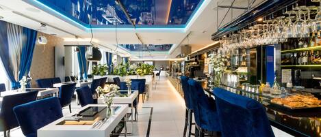 A modern restaurant with a high ceiling, blue lighting, and a bar area with wine glasses.