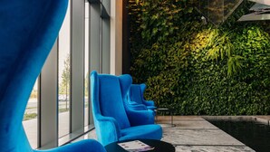 A seating area with blue chairs and a small table, a green wall with plants, and a view of the street through large windows. - Varsovia Comfort 223 (Warsaw)