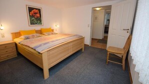A bedroom with a wooden bed, a chair, a nightstand, and a door leading to another room. - Apartment | 2 people (handicapped accessible) - main building (Carolinensiel)