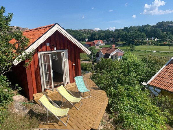 Una cabaña de madera con terraza y dos sillones de descanso, rodeada de vegetación y otras construcciones en un entorno rural.