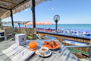 Un tavolo con un giornale, un croissant, una tazza di caffè e un bicchiere di succo d'arancia, con vista su una spiaggia con ombrelloni e sedie.