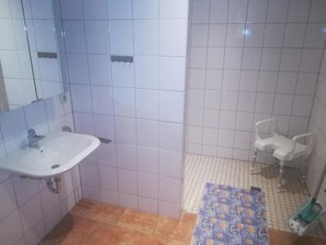 Une salle de bain avec des carreaux blancs, un lavabo, un miroir et un espace douche agrémenté d’une chaise blanche.