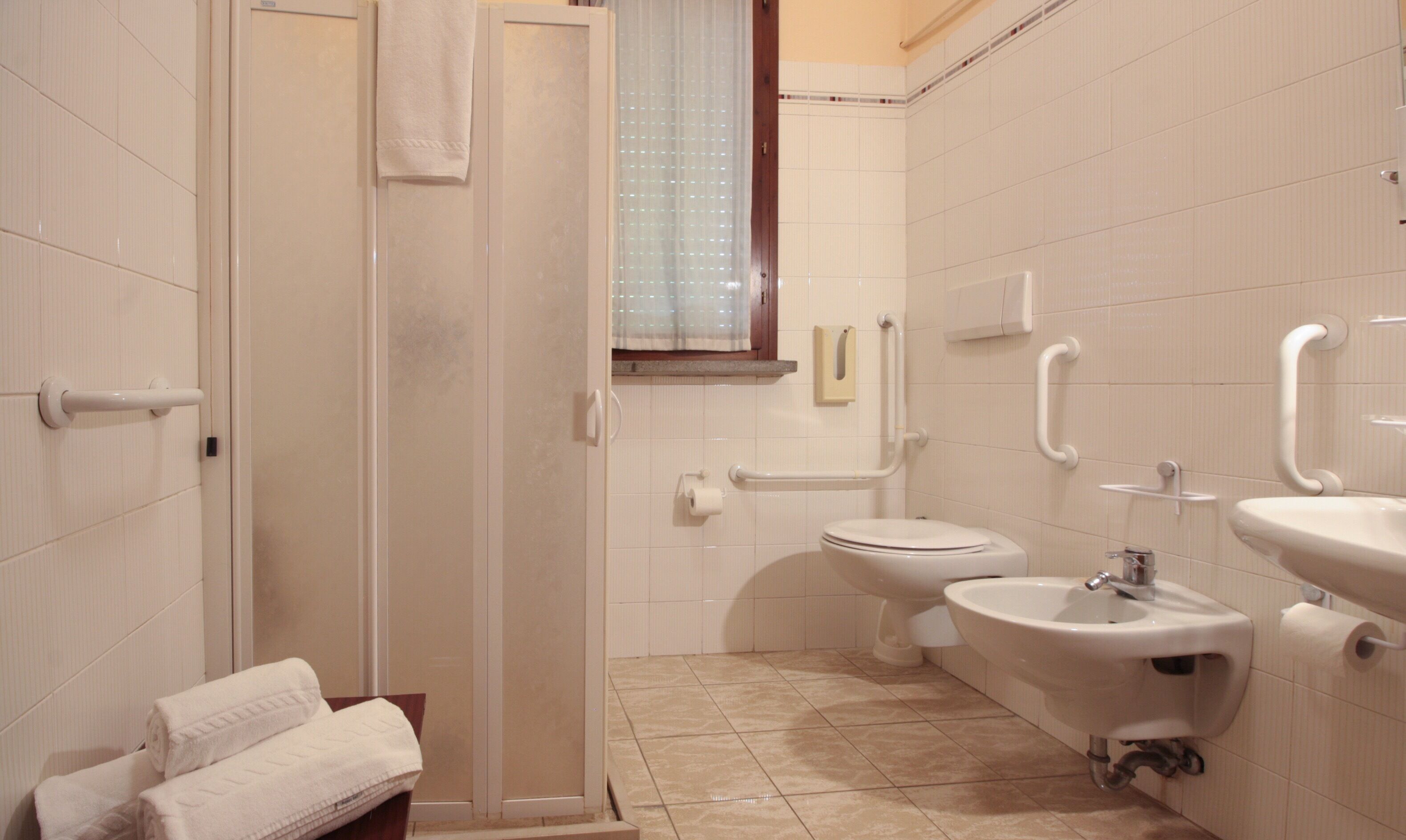 Baño con inodoro, bidé y lavamanos, equipado con barras de apoyo y zona de ducha.