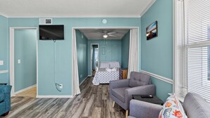 A living room with teal walls, a ceiling fan, a flat-screen TV, and a sofa. - Recently Renovated Oceanfront Condo in Windy Hill + Free Attraction Tickets! (North Myrtle Beach)