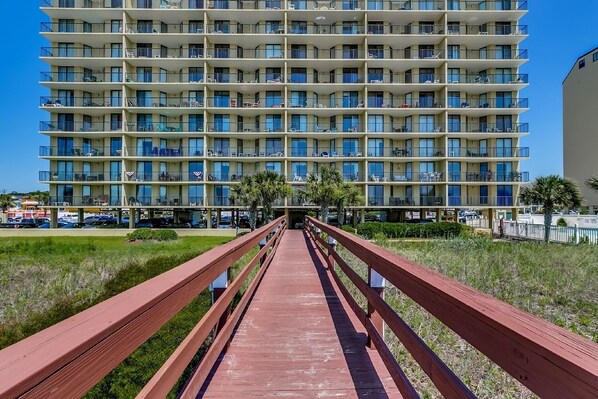 A multi-story hotel with balconies, a wooden walkway, and a parking area in the background.