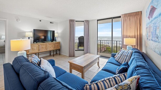 A modern living room with a large sofa set, a flat-screen TV, a wooden coffee table, and a balcony with a view of the sea.