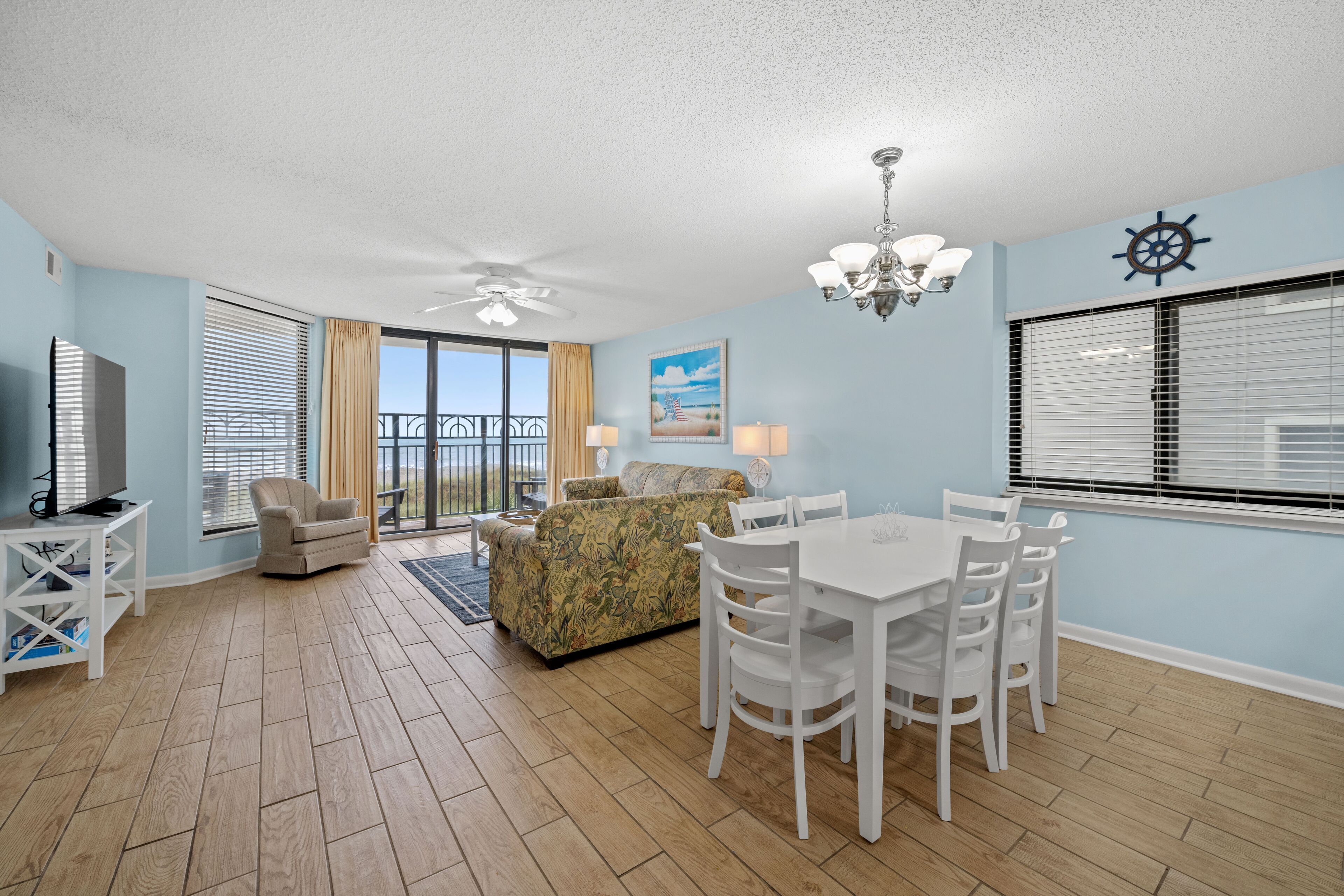 A spacious living room with a large window, a ceiling fan, a dining table with chairs, and a sofa.