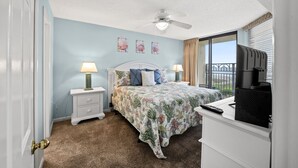 A bedroom with a bed, nightstands, a ceiling fan, a TV, and a window with blinds. - Spacious 1st floor corner Oceanfront unit + Free Attraction Tickets! (North Myrtle Beach)