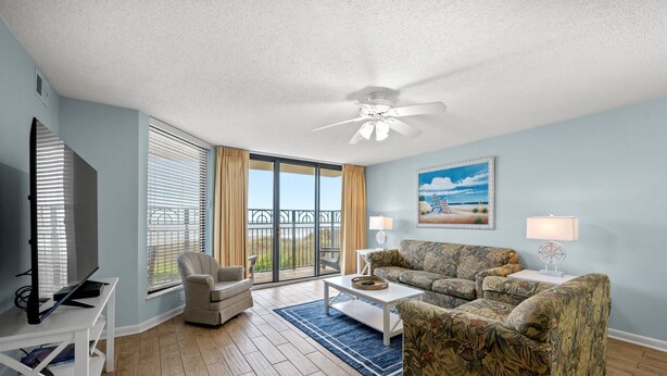 A living room with a ceiling fan, a television, a sofa, a chair, and a table. - Spacious 1st floor corner Oceanfront unit + Free Attraction Tickets! (North Myrtle Beach)