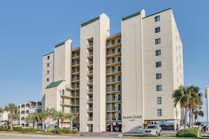 Un hotel de varios pisos con balcones, un letrero que dice "Beach Club" y un estacionamiento con coches.