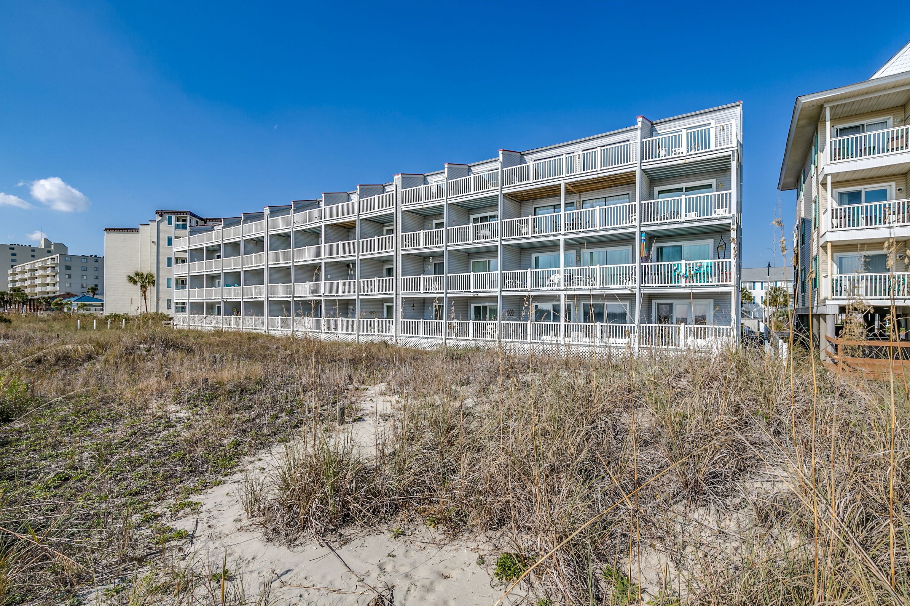 Ein Strandhotel mit mehreren Balkonen, ein Sandstrand davor und ein klarer, blauer Himmel.