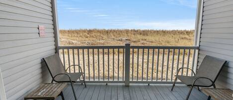 A covered porch with two wooden tables and four chairs, overlooking a sandy beach with tall grasses.
