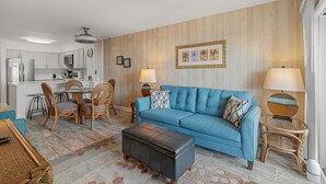 A modern living room with a blue sofa, a dining area with wicker chairs, and a kitchen in the background.