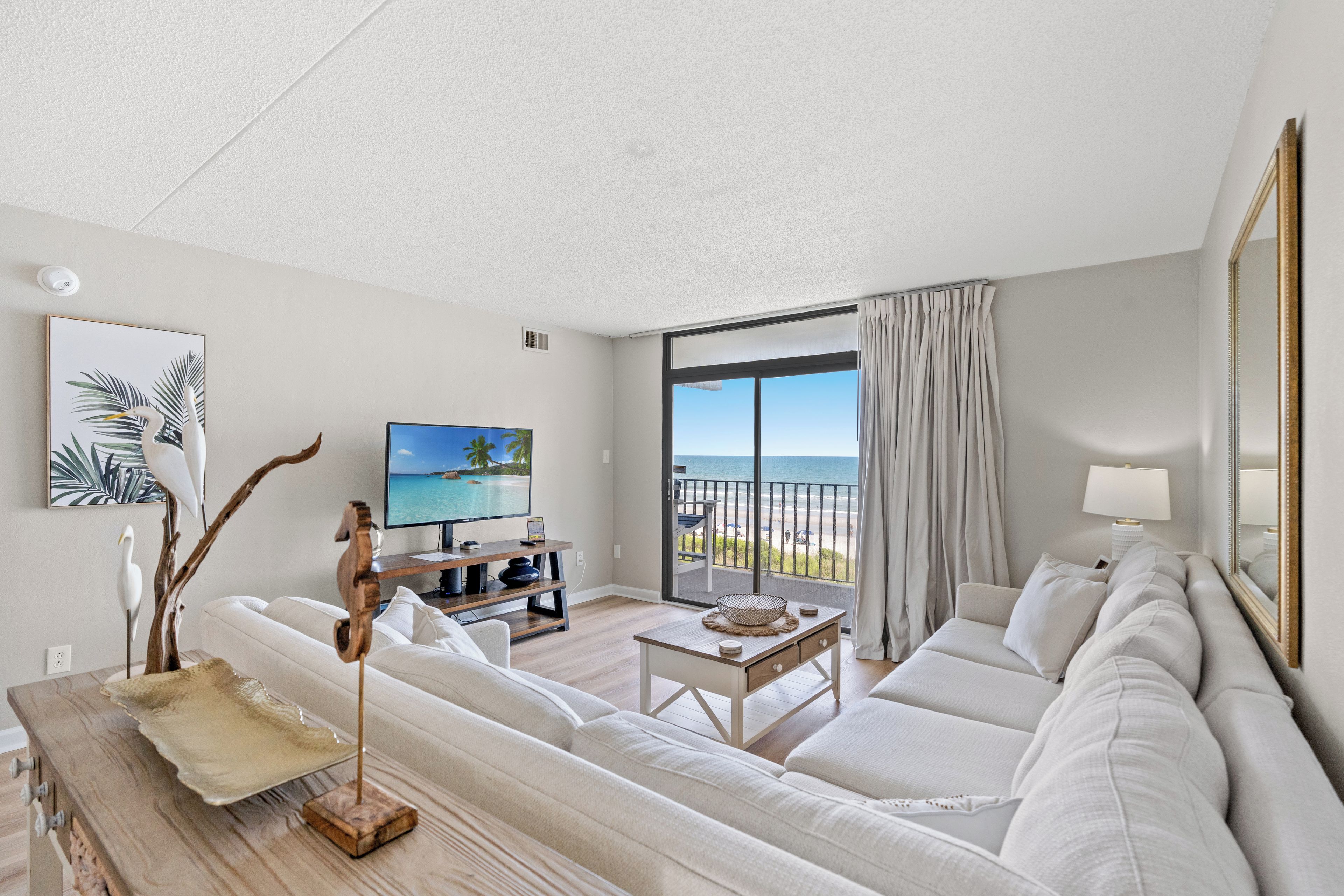 A modern living room with a sofa, a coffee table, and a television. There is a balcony with a view of the ocean.