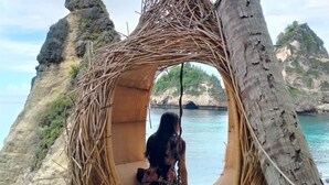 A woman sitting in a woven hanging chair overlooking a coastal area with rocky cliffs and clear blue water. - Ramp Cottage (Penida Island)