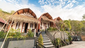 A bungalow-style accommodation with thatched roofs, a staircase leading to the entrance, and a garden area with various plants. - Ramp Cottage (Penida Island)