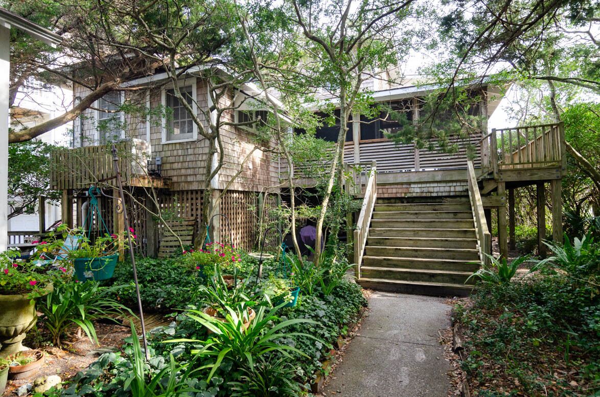 Un escalier en bois mène à une terrasse, qui est entourée d’une végétation luxuriante et d’une maison à la façade en bois.