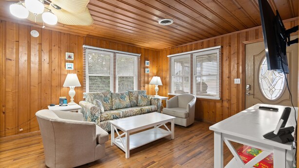 A room with wooden walls and ceiling, a sofa, two armchairs, a coffee table, a TV, and a door with a window.