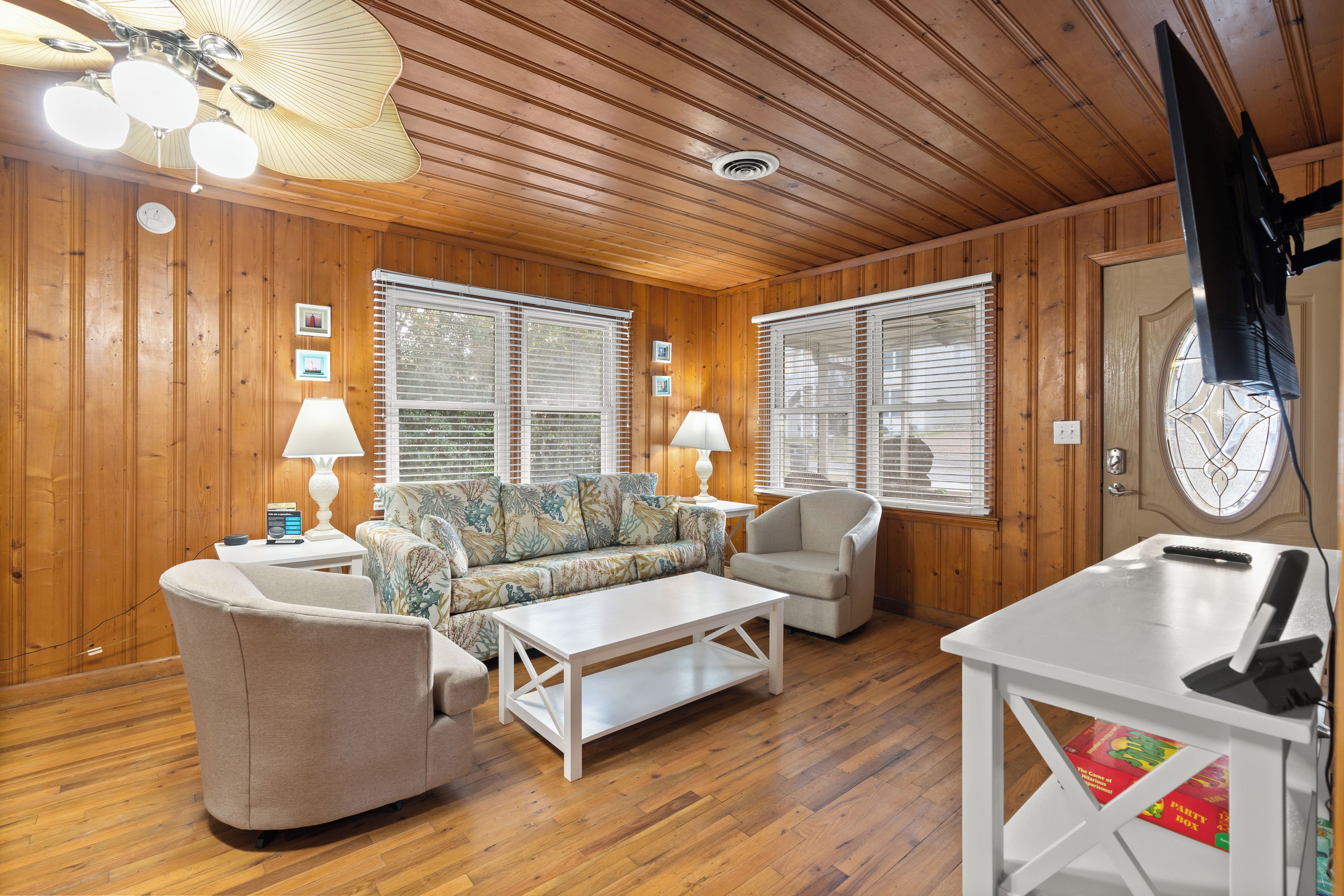 A room with wooden walls and ceiling, a sofa, two armchairs, a coffee table, a TV, and a door with a window.