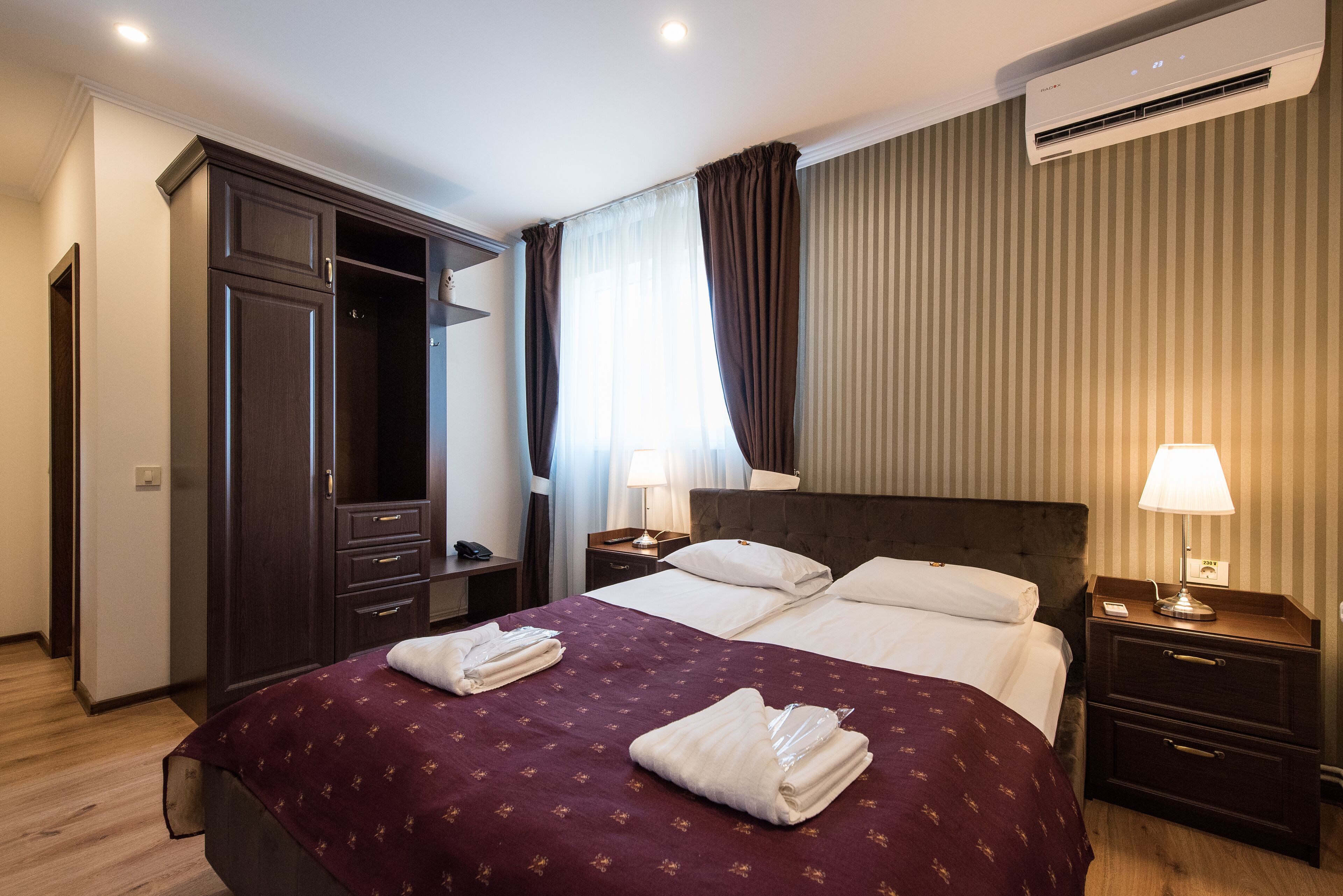 Una habitación de hotel con una cama grande, un armario, una mesita de noche con lámpara y una ventana con cortinas.
