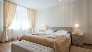 Un dormitorio ordenado con una cama grande, dos mesitas de noche con lámparas, un banco y una ventana con cortinas.