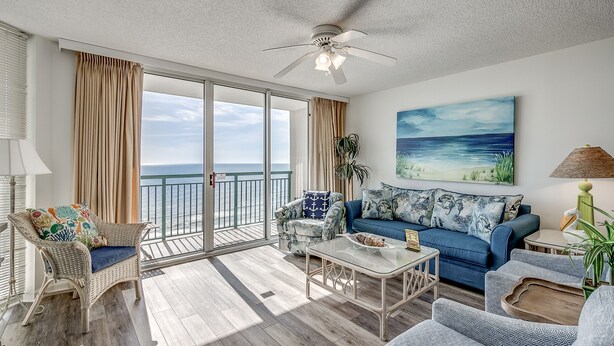 A spacious living room with a balcony, wicker furniture, a blue sofa, and a painting of a beach. - 10th floor newly remodeled unit + Free Attraction Tickets! (North Myrtle Beach)