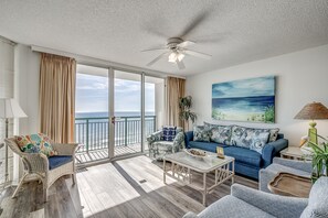 Ein geräumiges Wohnzimmer mit Balkon, Korbmöbeln, einem blauen Sofa und einem Strandbild.