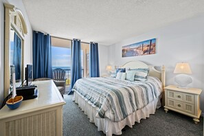 A hotel room with a bed, a desk, a chair, a nightstand, and a view of the ocean.